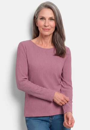 Femme aux longs cheveux gris portant un haut mauve texturé à manches longues et un jean bleu, debout devant un fond blanc.