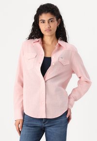 Camisa de botão rosa claro com mangas longas, dois bolsos frontais e um hem arredondado, feita de um tecido macio e texturizado. Apresenta botões brancos.