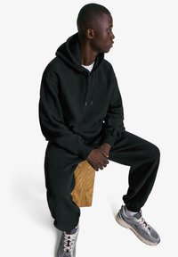 Čierna mikina s kapucňou a tepláky z mäkkej látky. Model sedí na drevenom stoličke, na nohách má šedé tenisky s červenými akcentmi. Jednoduchý, uvoľnený dizajn.