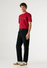 Rode katoenen t-shirt met korte mouwen en een borstzak; gecombineerd met zwarte ribfluwelen broek en witte sneakers met zwarte accenten.