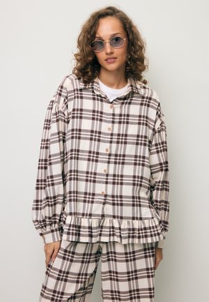 Vrouw met krullend haar, draagend een ronde zonnebril, een loszittend beige en bruin geruit overhemd met knopen, en bijpassende geruite broek.