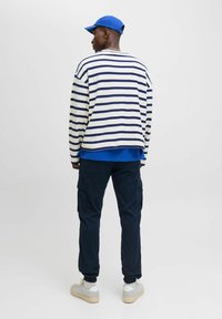 Langärmliges gestreiftes Hemd in Weiß und Navy, darüber ein blaues Oberteil. Kombiniert mit Cargo-Hosen in Navy und grauen Turnschuhen. Eine blaue Mütze akzentuiert das Outfit.