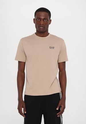 Beige bomulds t-shirt med rund hals og korte ærmer, prydet med et lille sort logo på venstre bryst. Glat tekstur, afslappet pasform.