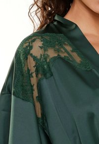 Vêtement en satin vert foncé avec des accents en dentelle verte intricats sur les épaules, créant une texture lisse avec un doux éclat.
