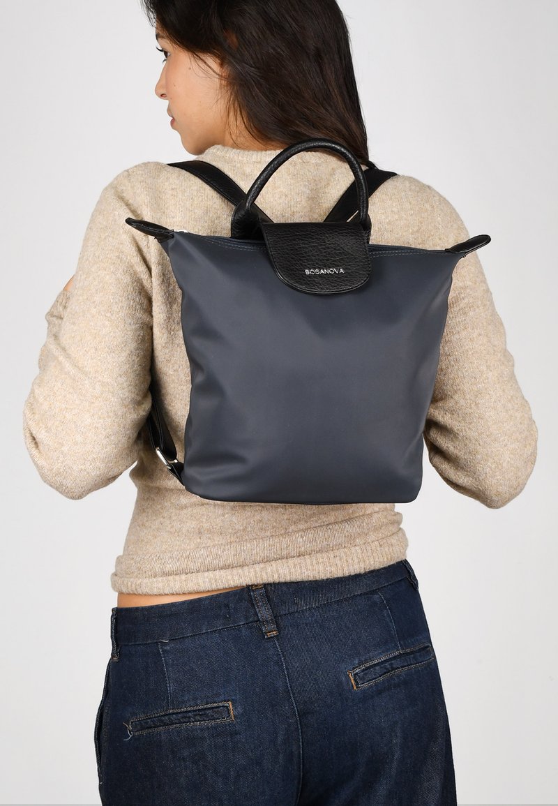 Mochila gris con detalles de cuero negro, con una tapa superior redondeada y dos correas ajustables, vista desde la parte trasera sobre una persona.