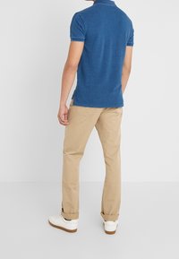 Man i blå kortärmad polo, beige byxor med uppvik, och vita sneakers, stående vänd mot en vit vägg, ryggen synlig.