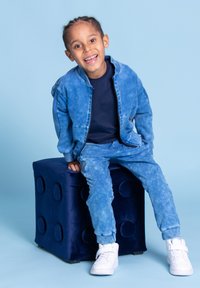 Niebieska kurtka dżinsowa i pasujące spodnie z wzorem tie-dye. Białe sneakersy, siedząca na miękkim niebieskim sześcianie. Jasnoniebieskie tło.