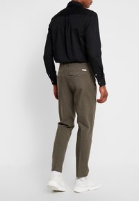 Černá košile s dlouhým rukávem a knoflíkovými manžetami, spárovaná s olivově zelenými kalhotami s kapsami na zadní straně. Boty jsou bílé sportovní tenisky.