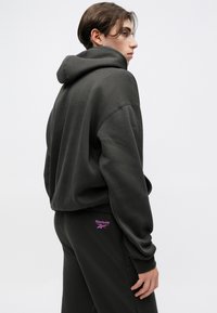 Tmavě šedá, oversized mikina s kapucí vyrobená z jemné látky. Na spodní levé straně se nachází malé fialové logo Reebok.