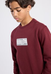 Maroon sweatshirt with a round neckline, featuring a rectangular gray patch with "THE FIVE BOROUGHS" and "NEW YORK" graphic detail.