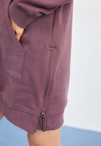 Lilla joggedress med glidelås på siden, ribbestrikkede mansjetter og en frontlomme. Stoffet ser mykt og teksturert ut.