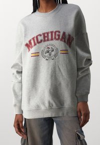 Šedá mikina s nápisem "MICHIGAN" v červených písmenech, která obsahuje grafickou pečeť a pruhy v žluté a červené barvě. Oversized střih s žebrovanými manžetami.