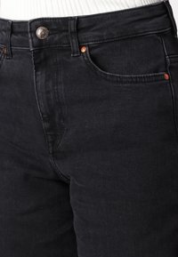 Gros plan sur un jean en denim noir montrant les passants de ceinture, les rivets en cuivre, la fermeture par bouton et la poche avant avec des détails de couture.