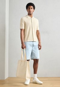 Beige textured knit polo shirt, light denim shorts, white sneakers, holding a beige tote bag with long straps. Neutral colors and casual design.