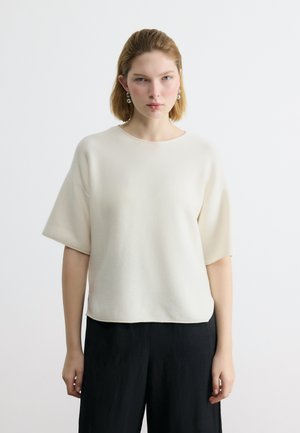 Noor naine lühikeste blondide juustega, kes kannab valget lühikese varrukaga topi ja musti pükse, seisab ühevärvilise valge taustaga ees.