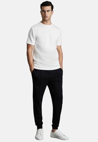 Camiseta blanca de manga corta combinada con pantalones jogger negros con costuras laterales y zapatillas blancas con cordones sobre un fondo liso.