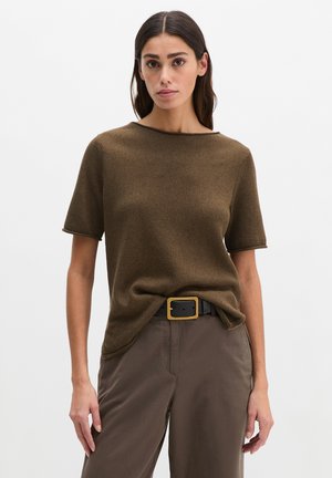 Femme portant un haut en maille à manches courtes marron rentré dans un pantalon marron avec une ceinture noire ornée d'une grande boucle dorée, debout devant un fond uni.