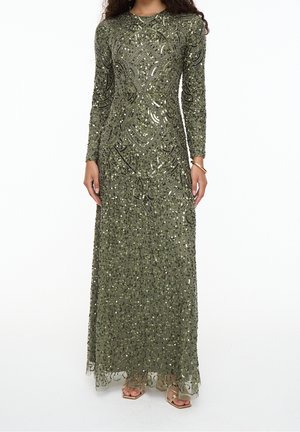 Longue robe vert olive avec broderies complexes de perles et paillettes, manches longues et col montant, portée avec des sandales à lanières dorées.