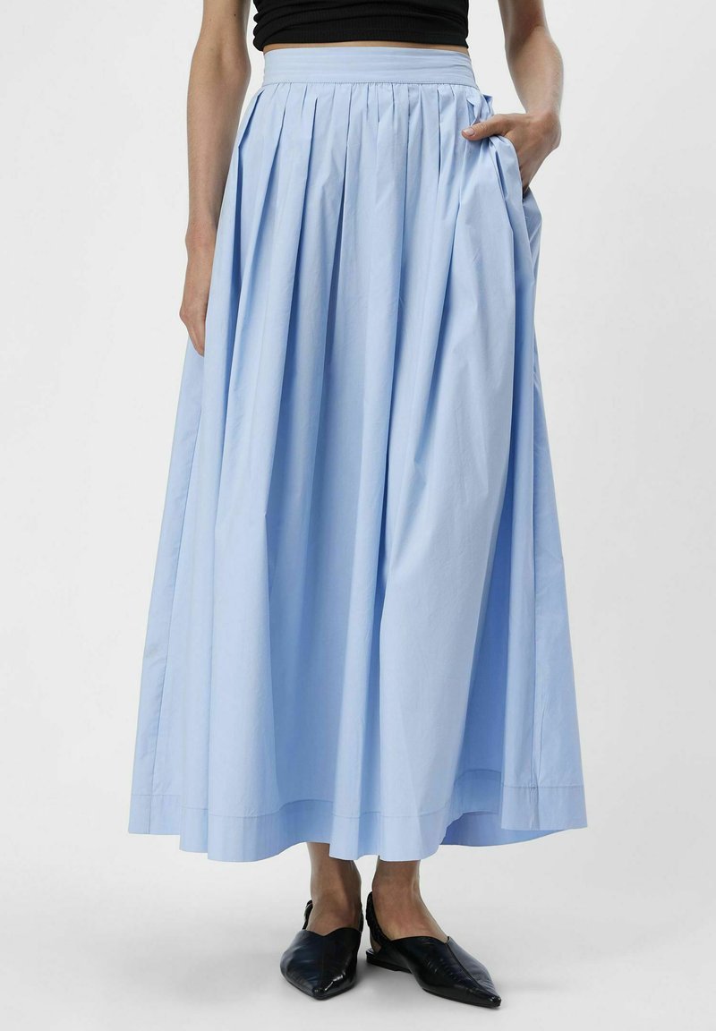 Jupe longue plissée bleu clair en tissu léger, dotée d'une large ceinture et de poches latérales. Portée avec des chaussures noires sans lacets.