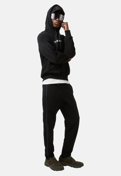 Sort hættejakke med logo foran, sorte sweatpants med sidelister, oversized spejlsolbriller og olivengrønne sneakers.