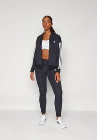 Casaco desportivo preto com riscas brancas, combinado com um top desportivo branco e leggings pretas. Tênis desportivos brancos completam o visual.