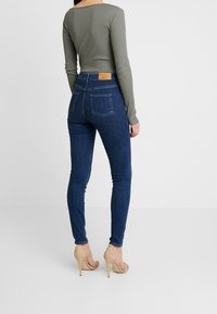 Smala jeans i mörkblå denim med en jämn yta, tillsammans med en figurnära olivgrön långärmad topp. Bakifrån syns två fickor och en etikett.