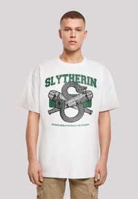 Bílý bavlněný tričko s grafikou Slytherin, která obsahuje design hada a kouzelné hůlky v zelené a šedé barvě. Krátké rukávy, kulatý výstřih. Volný střih.
