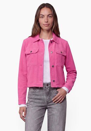 Femme portant une veste en velours côtelé rose vif sur une chemise blanche, associée à un jean gris, debout avec une main dans la poche.