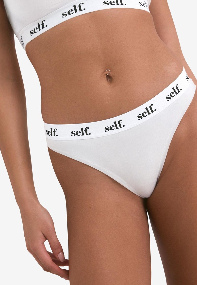 Biele tangá z mäkkej látky. Pás má opakujúce sa čierne logo "self.". Hladká textúra s dizajnom s minimálnym krytím.