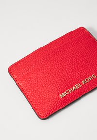 Raudona struktūrinė odinė kortelių laikiklis su auksiniu "MICHAEL KORS" logotipu, turintis tris kortelių vietas ir lygią juodą vidinę pamušalą.