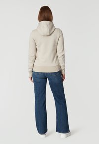 Beige hoodie med en dragsko huva, ribbade ärmslut och nederkant, kombinerad med mörkblå utsvängda jeans och vita sneakers, sedd från baksidan.
