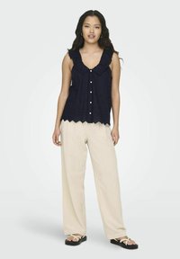 Marineblauwe mouwloze blouse met gerimpelde kraag en golvende zoom, gesloten met witte knopen. Gepaard met losse beige broek en zwarte sandalen.