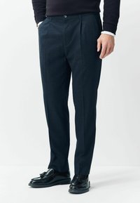 Dark blue dress pants with a textured finish, featuring pleats, a button closure, and side pockets, paired with black loafers.