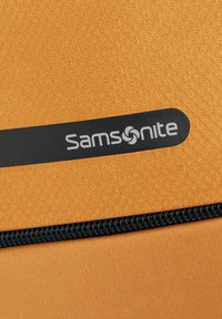 Tessuto arancione testurizzato con una cerniera nera. Presenta un'etichetta nera con il logo bianco "Samsonite" ben visibile.