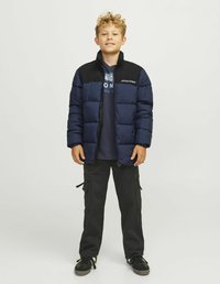 Marinblå isolerad pufferjacka med svarta accenter, med hög krage och framsidan dragkedja. Bärs över en marinblå grafisk t-shirt och svarta cargo-byxor.
