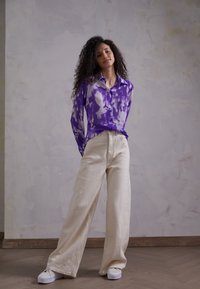 Camisa de botão em tie-dye roxo e creme com mangas longas, combinada com calças largas creme e sapatos brancos sobre um fundo texturizado.