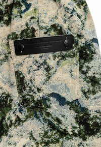 Tejido texturizado con un patrón abstracto en verde, azul y negro sobre un fondo beige, con una etiqueta rectangular negra que muestra los detalles de la marca.