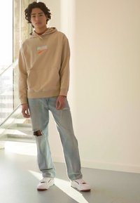 Beige hettegenser med lommer foran og PUMA-logo, kombinert med lyseblå slitte jeans og hvite sneakers med røde detaljer.