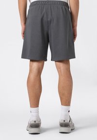 Uomo con pantaloncini grigio scuro, calze atletiche bianche e sneakers grigie, visto da dietro su uno sfondo chiaro e uniforme.