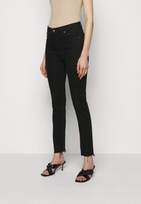 Kvinna iförd svarta högmidjade slim jeans med fransade kanter och svarta klacksandaler med flätade remmar, beige ärmlös topp synlig.