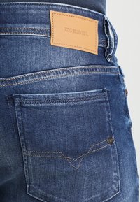 Mörkblå denimjeans har en läderlappar som är märkt "DIESEL", synlig sömnad och en karakteristisk design på bakfickan med ett litet märke.