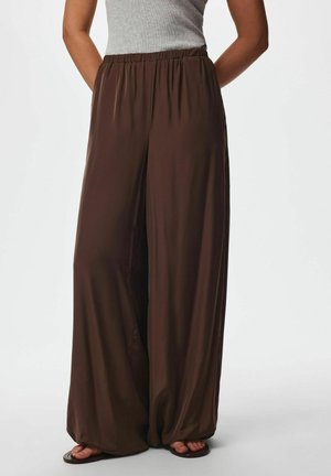 Personne portant un pantalon marron ample à jambes larges avec une taille élastique et des sandales marron type tongs, debout devant un fond uni.