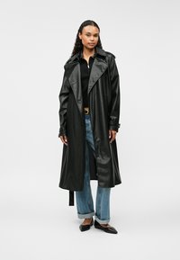 Czarny płaszcz trench z sztucznej skóry z szerokim kołnierzem, długimi rękawami i paskiem w talii. Noszony na czarną bluzkę i jasnoniebieskie dżinsy.