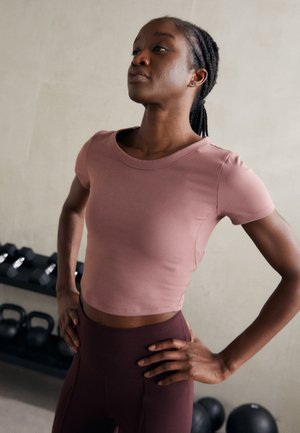 Atletski crop top od mekog ružičastog materijala s okruglim izrezom i kratkim rukavima, uparen s tajicama visokog struka u tamnoj burgundskoj boji.
