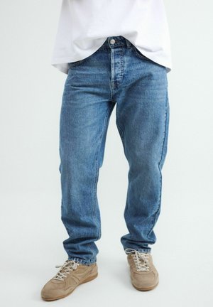 Osoba ubrana w niebieskie dżinsy i beżowe sneakersy stoi na jednolitym jasnym tle.