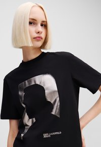 Melna T-kreklu ar metālisku sudraba siluetu un tekstu "KARL LAGERFELD JEANS" uzrakstā. Īsas piedurknes, apaļa kakla izgriezums.