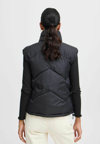 Czarna kamizelka puchowa z wysokim kołnierzem, chevronowymi przeszyciami i gładką, błyszczącą teksturą. Noszona na dopasowanej czarnej bluzce z długim rękawem.