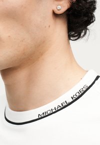 Κοντινή λήψη ενός λαιμού και αυτιού ατόμου που φοράει σκουλαρίκι με διαμάντι και λευκό πουκάμισο με το κείμενο "MICHAEL KORS" στον γιακά.