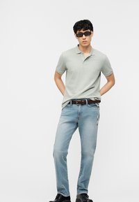 Νέος άνδρας με μαύρα γυαλιά ηλίου, ανοιχτό πράσινο πόλο μπλούζα, καφέ πλεκτή ζώνη, ανοιχτό μπλε τζιν και μαύρα παπούτσια, στέκεται με τα χέρια πίσω από την πλάτη.