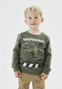 Zelená bavlnená mikina so grafikou dinosaura, textom "POZOR" a horizontálnymi pruhmi na spodnej strane. Dlhé rukávy a okrúhly výstrih.